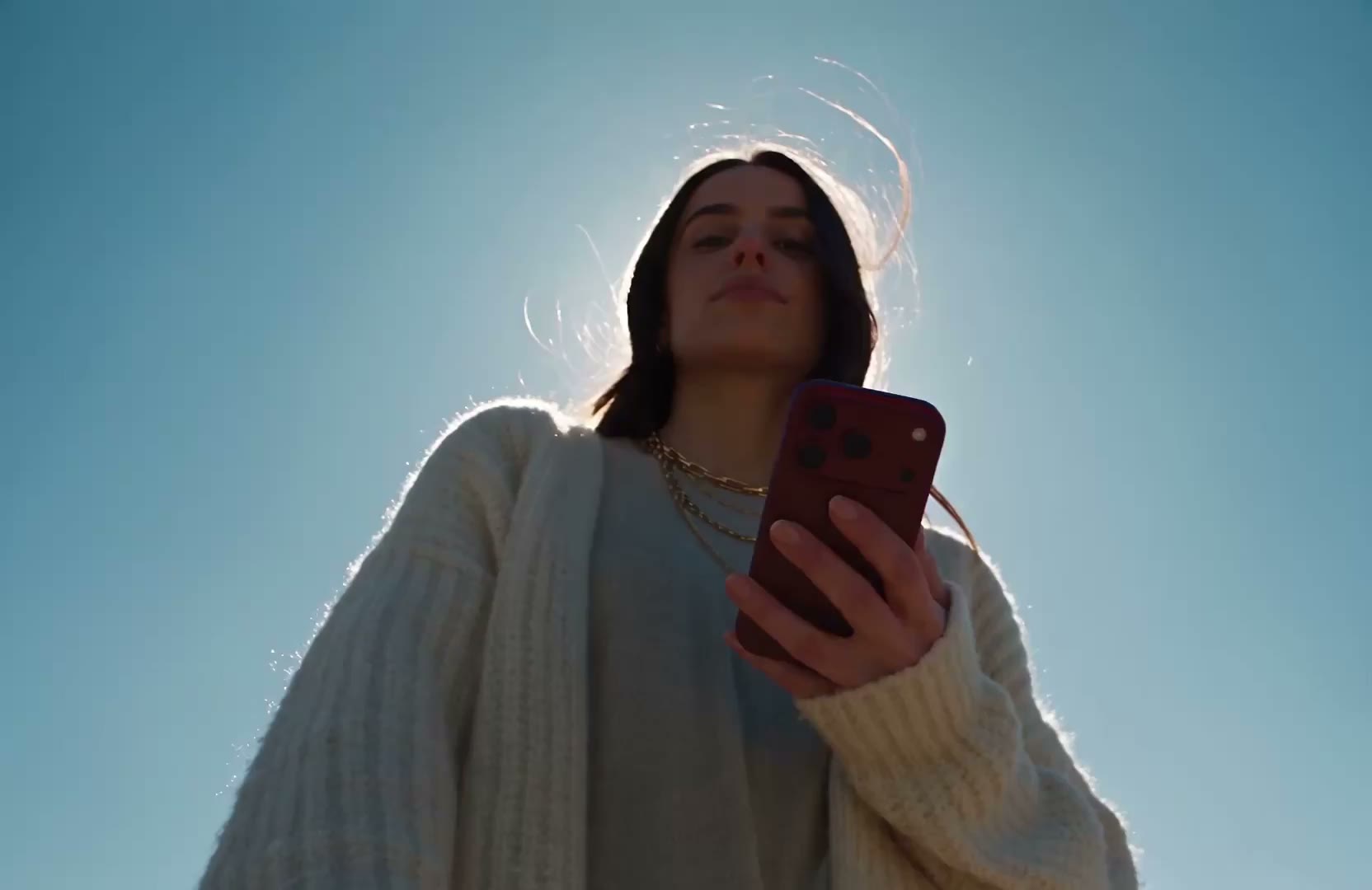 A woman in a white cardigan, seen from below against a bright blue sky, holding her phone in one hand.